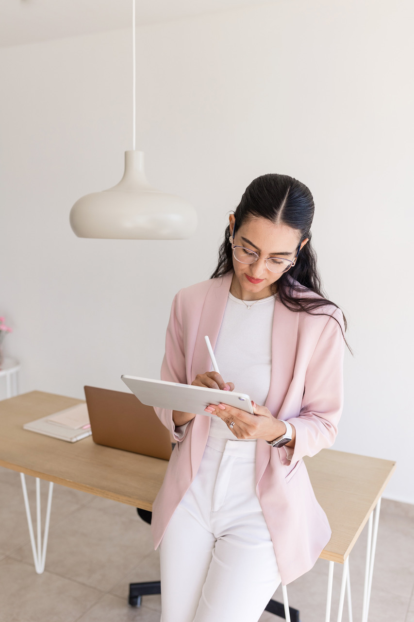 Girl is taking charge and writes ideas on a white tablet. She’s dressed in white trousers,white shirt, and pink jacket. She leans on a light brown desk in an office.