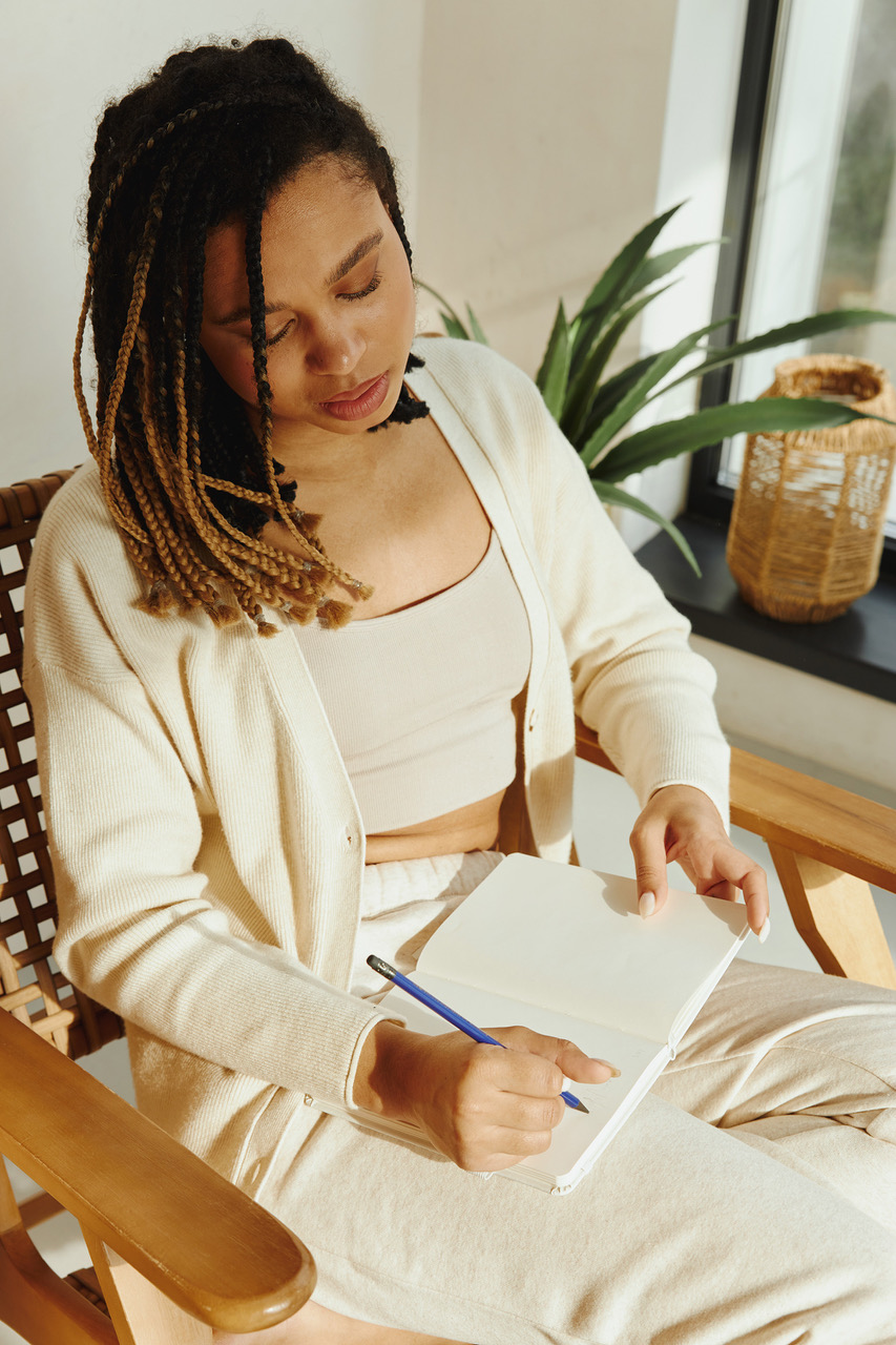 Woman of colour sits on a chair. She’s dressed in white lounge wear and writes in a journal to make decisions easier with decluttering the mind.