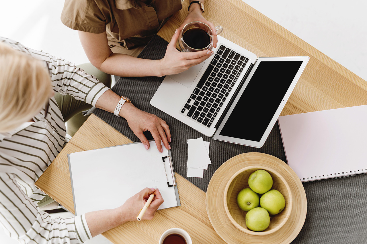 zwei Frauen an einem Arbeitstisch. Am Tisch stehen grüne Äpfel in einer Schüssel und Kaffeetassen. Es steht ein Laptop drauf und eine Frau schreibt beim Coaching auf ein Clipboard, denn echte Veränderungen brauchen neben positiven Gedanken auch aktive Handlungen.